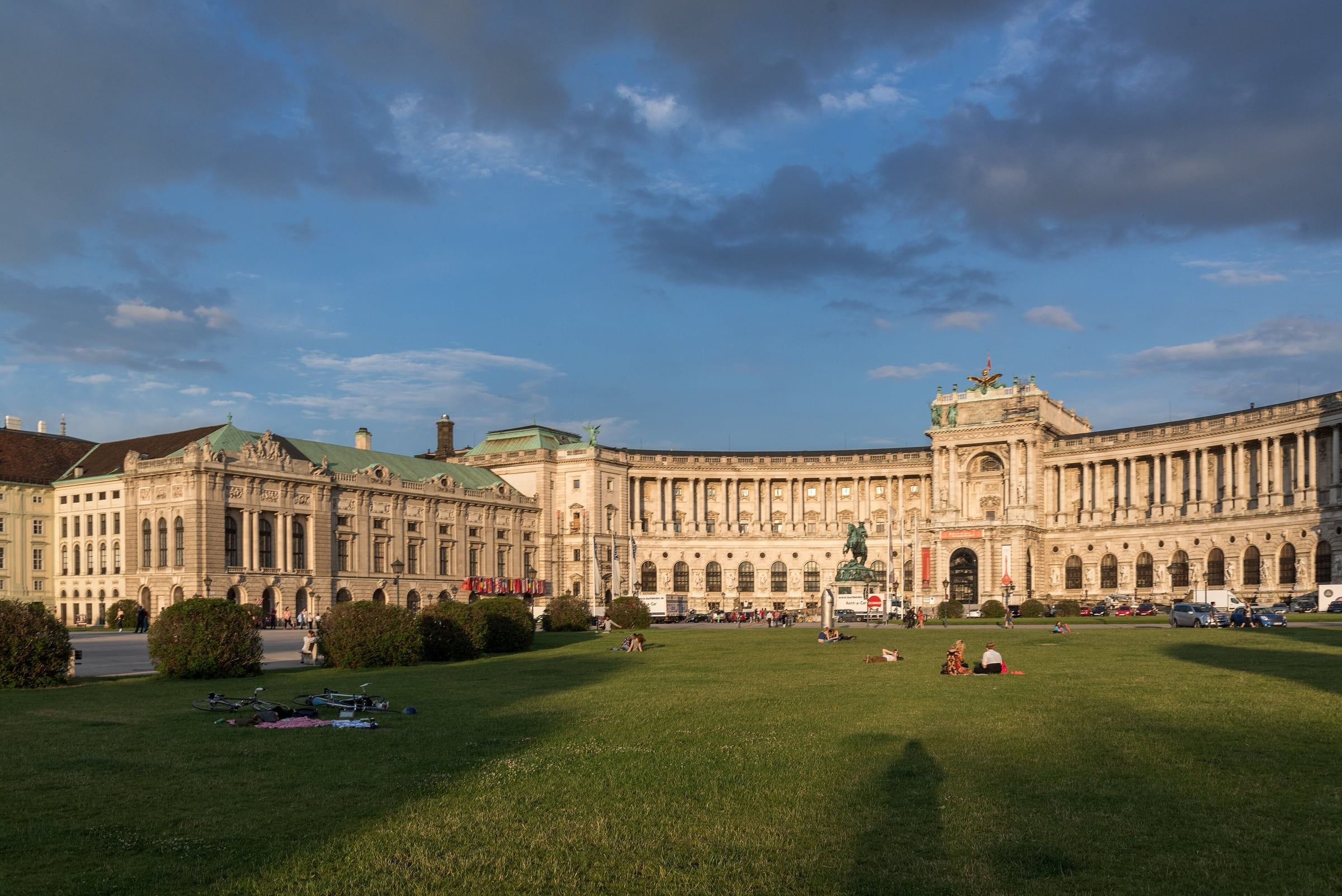 The curved Neue Burg facade of the Hofburg seen across the grassy Heldenplatz in warm evening light, with people relaxing on the lawn.