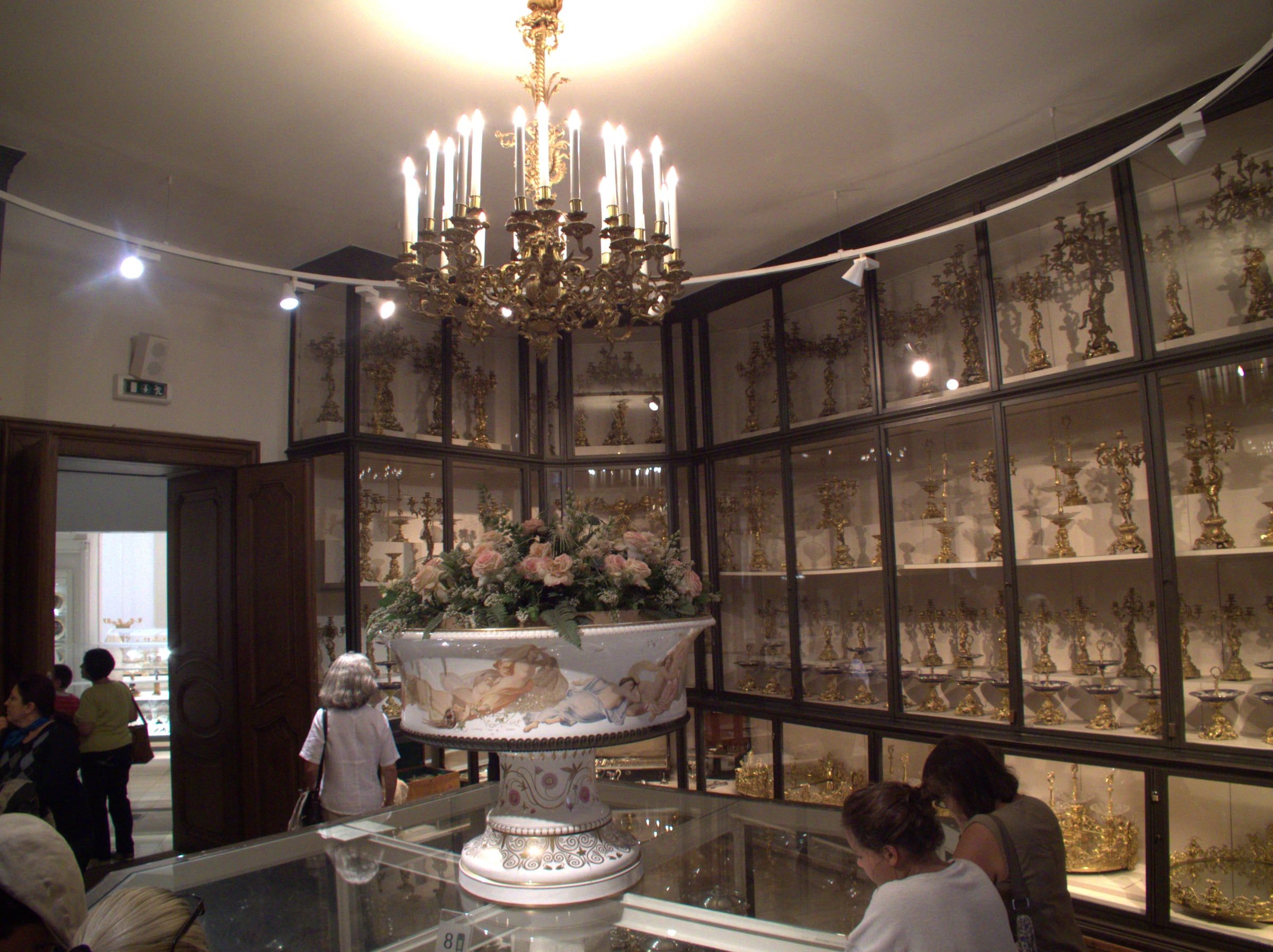 Ornate candelabra and gilt silverware fill glass cabinets beneath a gilded chandelier, with visitors and a painted porcelain centrepiece in front.