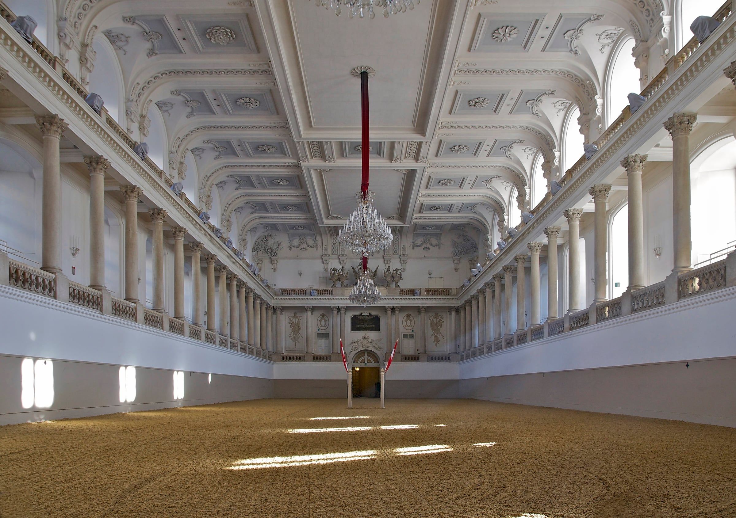 The empty Winter Riding School hall with its white Baroque galleries, columns, coffered ceiling and crystal chandeliers above the sand arena.